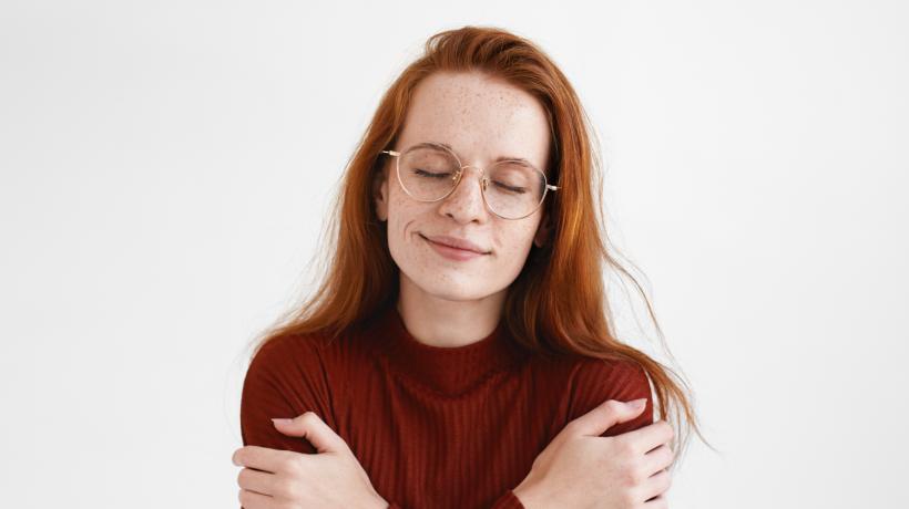 Eine Frau mit Brille und rötlichen Haaren umarmt sich selbst.
