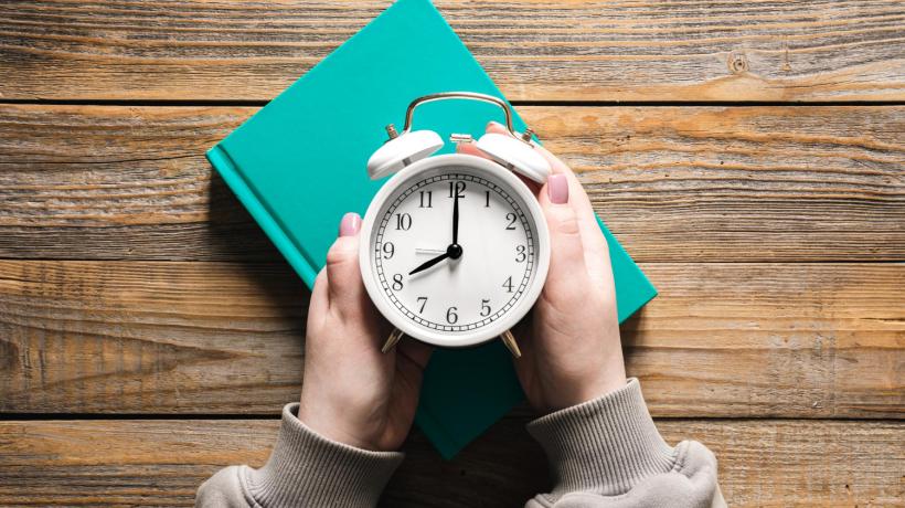 Man sieht einen weißen Wecker von oben, der von zwei Händen umfasst wird.