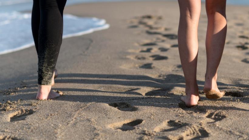 Man sieht die Beine zweier Personen, die barfuß durch den nassen Sand am Strand laufen.