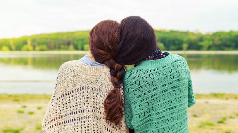 Man sieht zwei Frauen von hinten, die aneinandergeschmiegt auf einen See schauen.