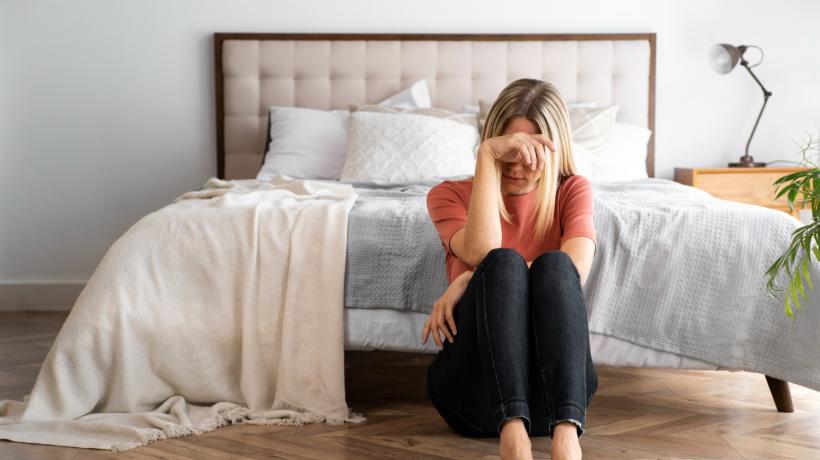Eine Frau kauert mit der Hand im Gesicht auf dem Boden vor ihrem Bett.