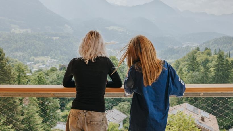 Zwei Jugendliche von hinten, die an eine Brüstung gelehnt in die Berge schauen.