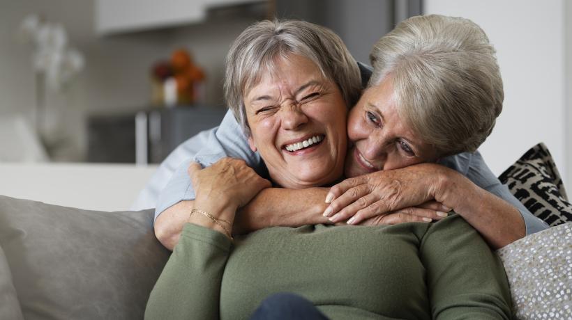 Eine ältere Frau umarmt eine etwa gleichaltrige Frau von hinten, beide lachen.