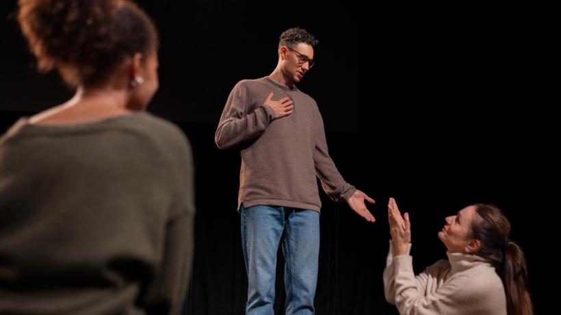 Ein Mann vor schwarzem Hintergrund, vor ihm kniet eine Frau, eine weitere Frau steht mit dem Rücken zur Kamera