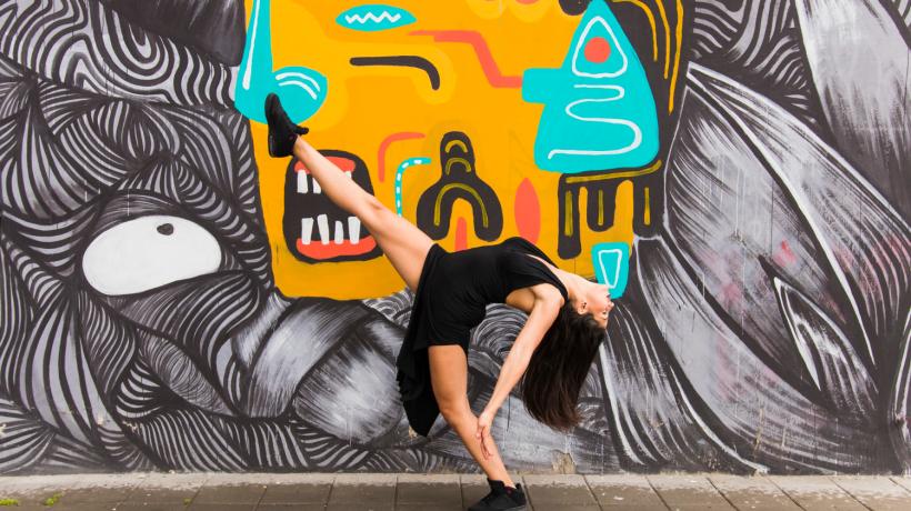 Eine Frau nimmt vor einer bunt bemalten Wand eine tänzerische Pose ein.