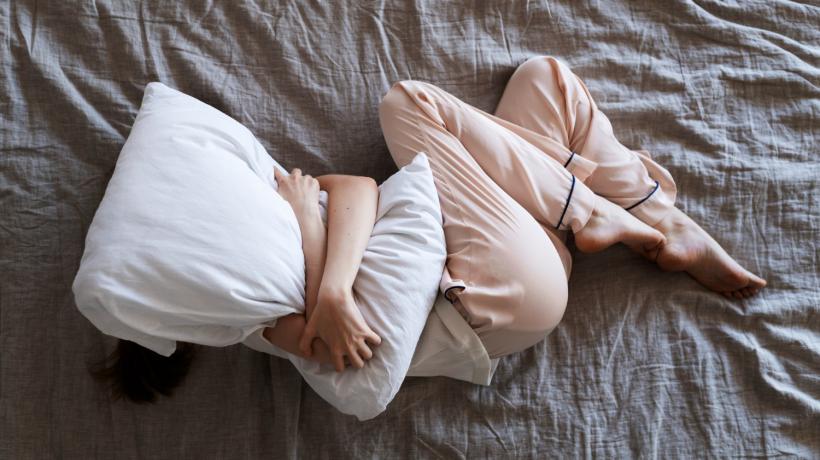 Man sieht eine Frau, die ein großes Kissen umarmt, auf einem Bett liegen. Ihr Gesicht ist unter dem Kissen veborgen.