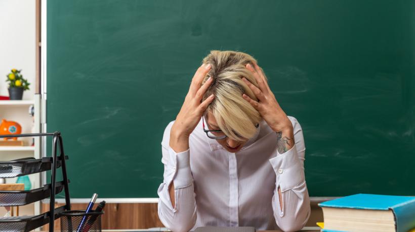 Frau am Lehrerpult, die ihren Kopf in die Hände stützt.