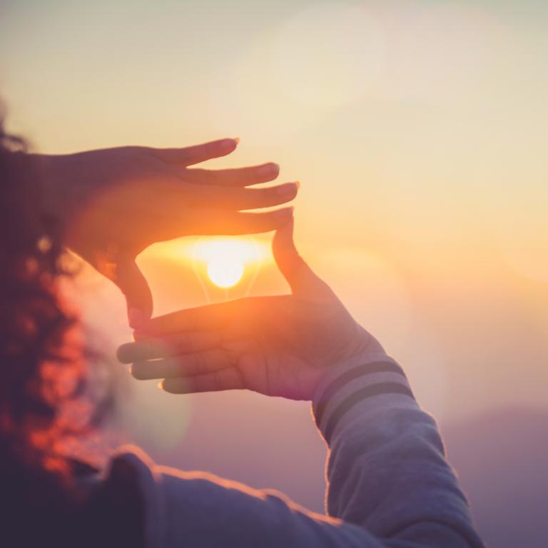Frau von hinten, die einen Sonnenuntergang beobachtet und mit Ihren Fingern ein Rechteck um die Sonne am Horizont formt.