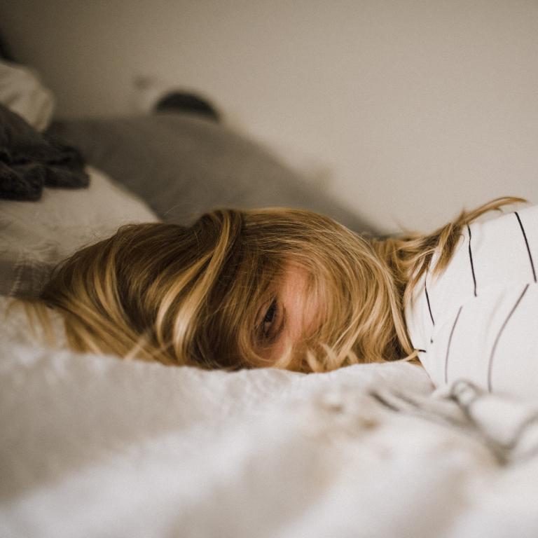 Frau liegt mit zerzausten Haaren auf dem Bauch auf einem Bett und sieht müde aus.