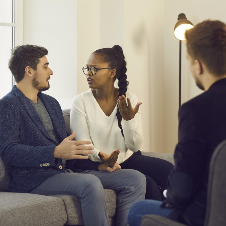 Therapieszene, ein Paar sitzt auf der Couch, spricht miteinander. Ihnen gegenüber sitzt ein anderer Mann, den man nur von hinten sieht.