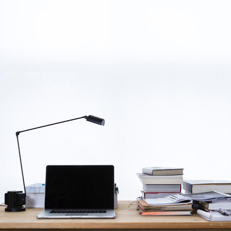 Frontansicht eines Schreibtischs mit Lampe, Laptop, Büchern und Dokumenten. Im Hintergrund eine weiße Wand oder ein Fenster mit sehr hellen Gardinen.