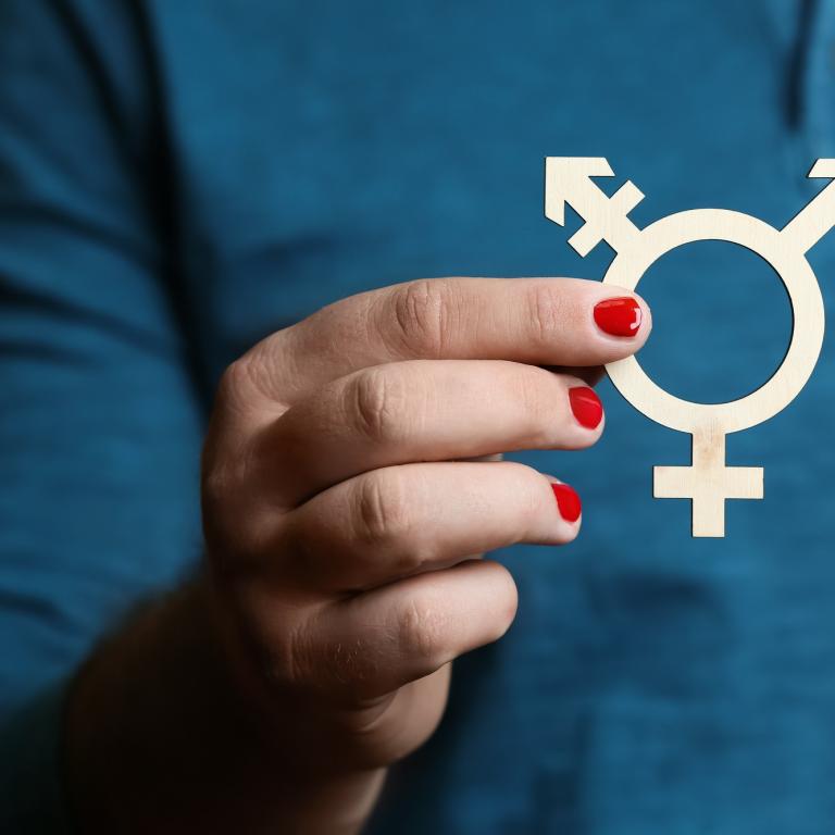 Ein Mann mit rot lackierten Fingernägeln hält das Transgender-Symbol vor sich.