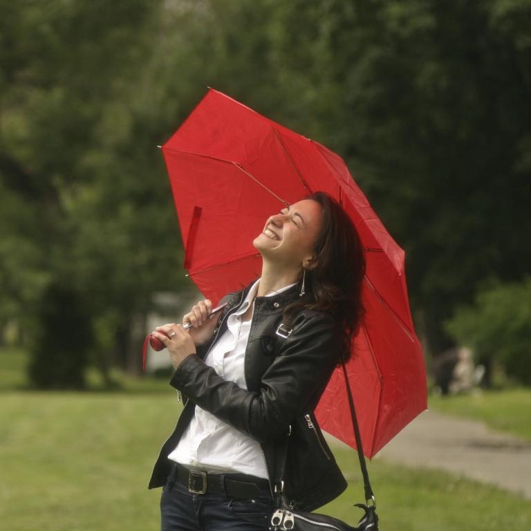 Eine weiße Frau steht lachend auf einer Wiese. Sie hält sich einen geöffneten roten Schirm über den Kopf. Im Hintergrund ein Baum.