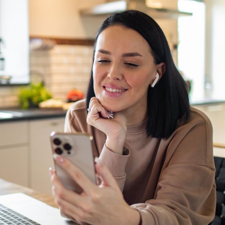 Eine Frau sitzt am Küchentisch, in der Hand ein Smartphone. Auf dem Tisch steht ein aufgeklappter Laptop.