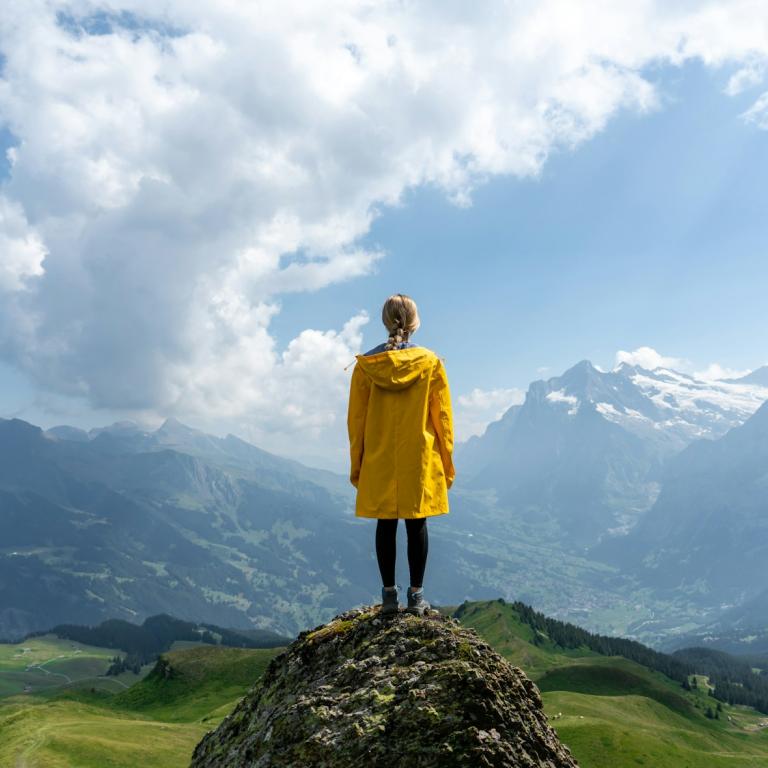 Rückansicht einer Frau in gelber Jacke, die von einer Anhöhe in eine bergige Landschaft schaut