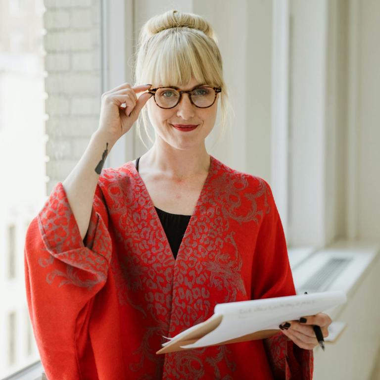 Eine blonde Frau mit Brille fasst sich an die Brille, sie hält einen Papierblock in der Hand.