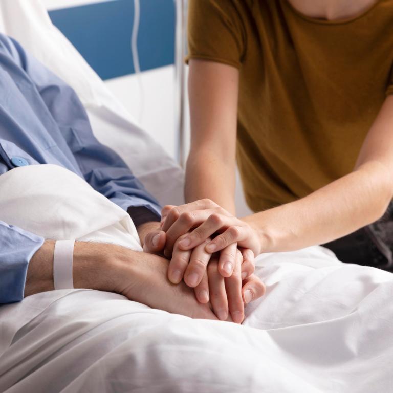 Eine junge Person hält ihre Hände auf die verschränkten Hände einer alten Person, die in einem Bett liegt.
