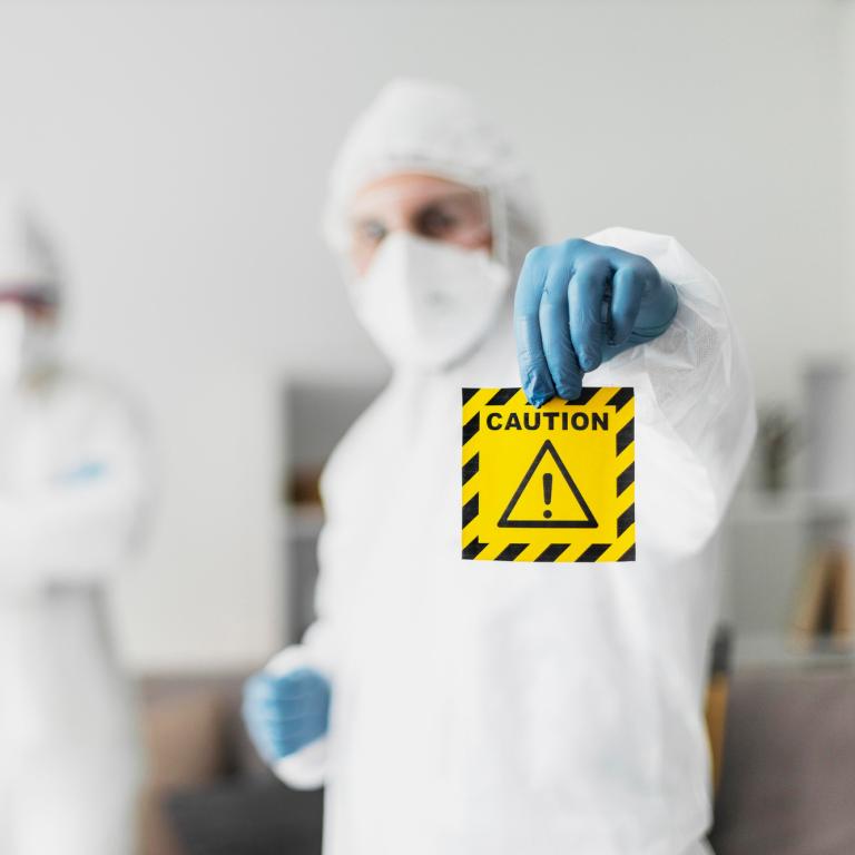 Zwei Personen in Hygieneschutzkleidung. Eine Person hält ein Achtung-Symbol hoch.