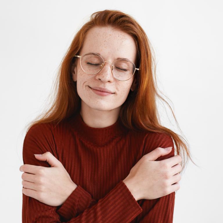 Eine Frau mit Brille und rötlichen Haaren umarmt sich selbst.