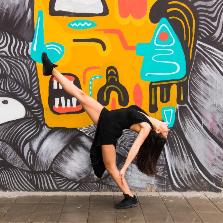 Eine Frau nimmt vor einer bunt bemalten Wand eine tänzerische Pose ein.
