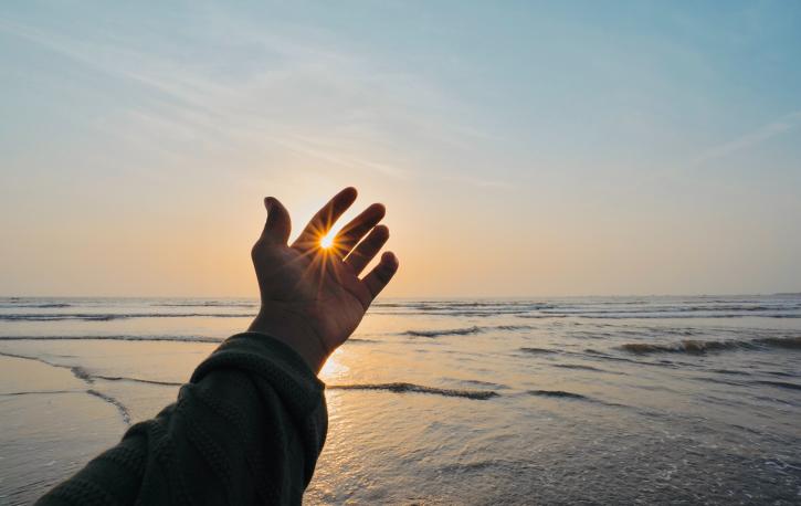 Hand vor Meer im Hintergrund, Sonnenstrahlen zwischen den Fingern sichtbar