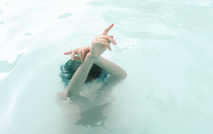 Frau unter Wasser bis zur Stirn. Eine Hand aus dem Wasser heraus in die Höhe gestreckt.