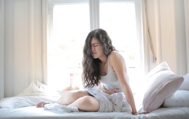 (Foto: Andrea Piacquadio – Pexels.com) Junge Frau sitzt im Bett und hält sich mit schmerverzerrtem Gesicht den Bauch.