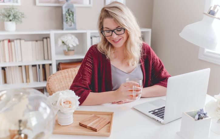(Foto: Paige Cody – Unsplash.com) Blonde Frau sitzt lächelnd am Schreibtisch vor aufgeklapptem Laptop.