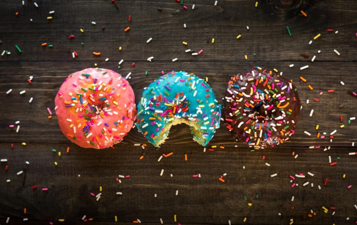Ein rosa, ein blauer und ein brauner Donut mit bunten Streuseln liegen auf einer dunklen Tischplatte, der mittlere Donut ist angebissen.