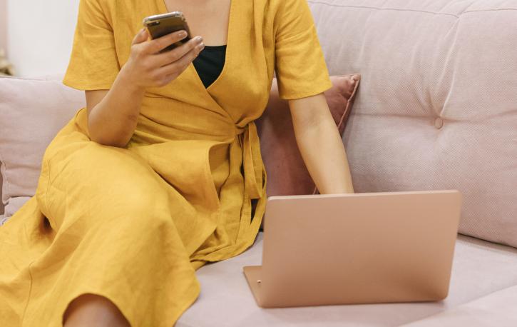 Frau in gelbem Kleid sitzt vor Laptop mit Handy in der Hand.