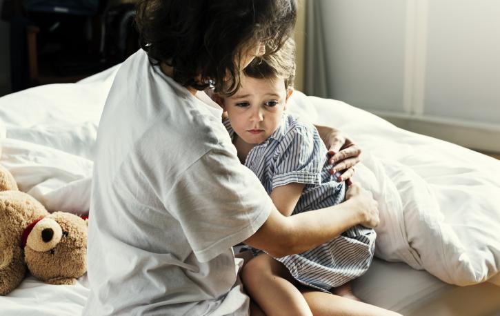 Mutter umarmt ihren kleinen Sohn, der ängstlich guckt. Beide sitzen im Schalanzug auf dem Bett.