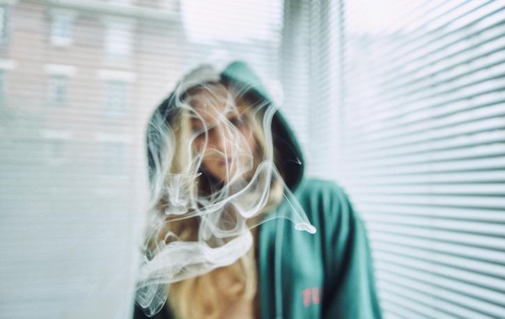 Eine Frau mit langen blonden Haaren und Kapuzenpulli steht vor halbgeschlossenen Jalousien am Fenster. Vor ihrem Gesicht qualmt Rauch.