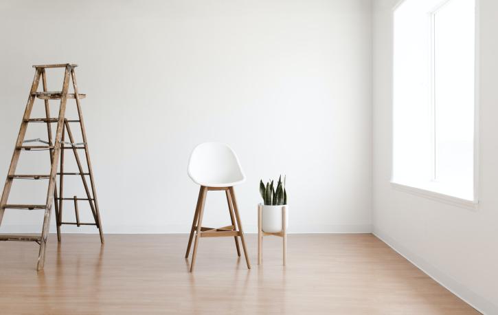 In einem leeren Raum vor einer weißen Wand stehen eine Leiter, ein Stuhl und eine Pflanze.