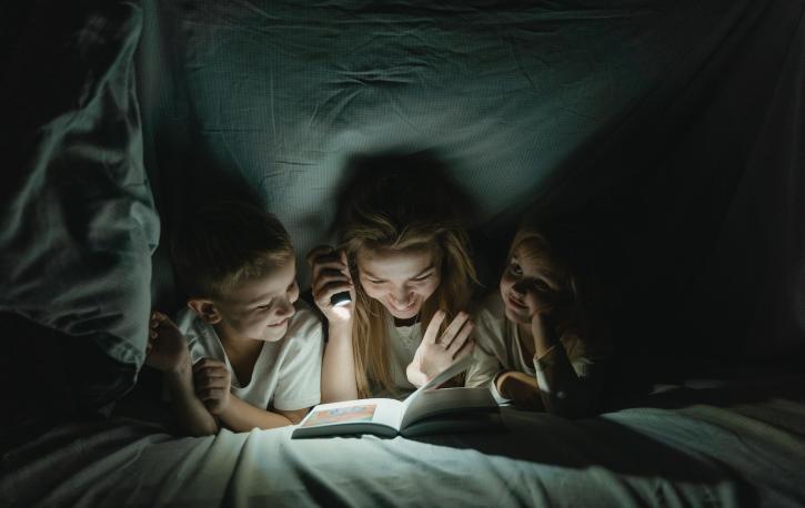 Eine Frau sitzt mit einem kleinen Jungen und Mädchen samt Taschenlampe unter der Bettdecke und liest ihnen aus einem Buch vor.