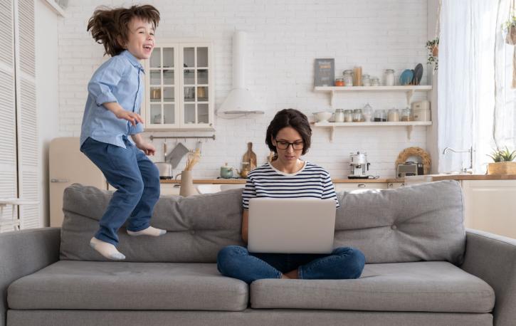 Eine weiße Frau sitzt im Schneidersitz und mit Laptop auf den Beinen auf einem grauen Sofa. Neben ihr springt ein kleines Kind auf dem Sofa auf und ab.