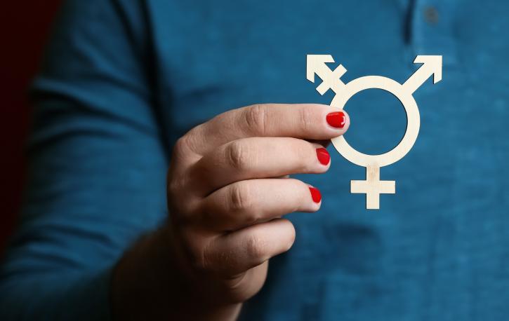 Ein Mann mit rot lackierten Fingernägeln hält das Transgender-Symbol vor sich.
