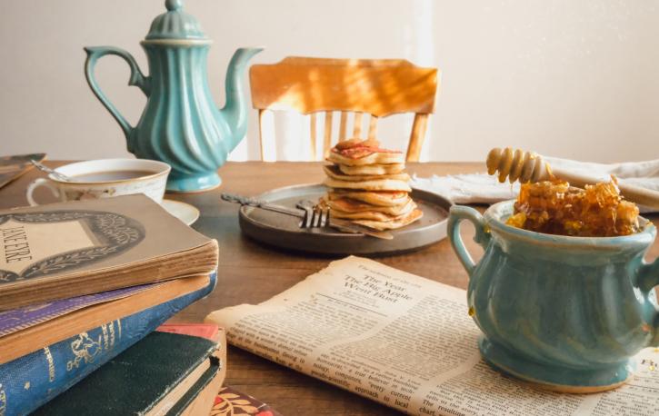 Auf einem Tisch steht ein Teller mit Pancakes, daneben eine Kanne Kaffe, ein Stapel Bücher und eine Zeitung.