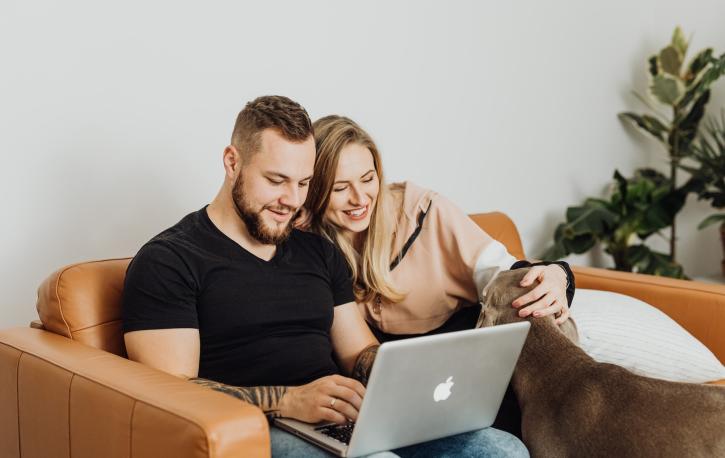 Ein Paar bestehend aus Mann und Frau sitzt auf der Couch und blickt auf einen Laptop, den er auf den Knien hält.