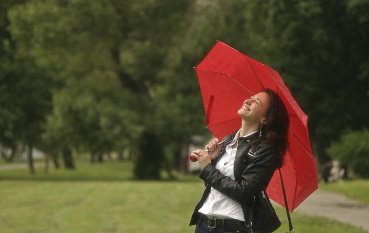 Eine weiße Frau steht lachend auf einer Wiese. Sie hält sich einen geöffneten roten Schirm über den Kopf. Im Hintergrund ein Baum.
