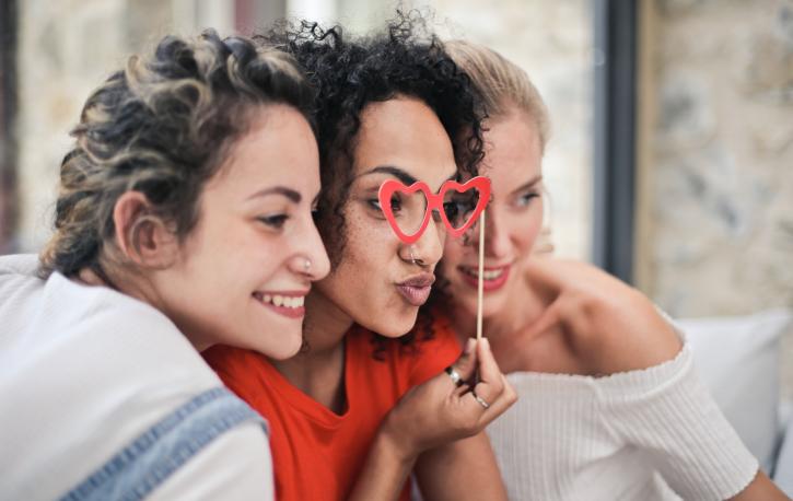 Drei Frauen, die ihre Köpfe nebeneinanderhalten, die mittlere Frau schaut durch eine rote Herzbrille