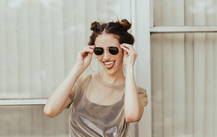 Eine junge Frau in einem dünnen Top und mit zwei Knubbelzöpfen greift lächelnd nach ihrer Sonnenbrille.