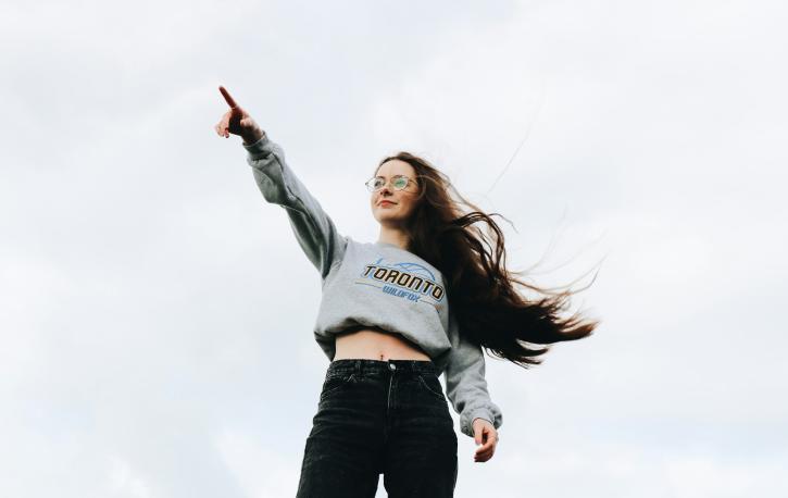 Eine junge Frau zeigt selbstbewusst in die Ferne, im Hintergrund ein bewölkter Himmel, ihre Haare flattern im Wind.