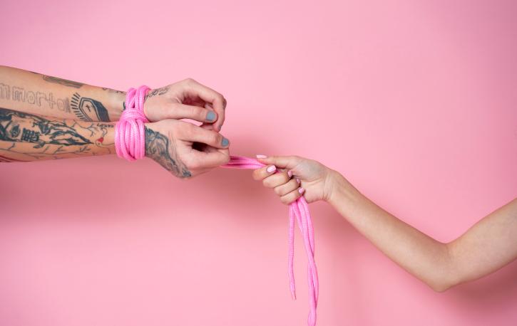 Vor einem rosa Hintergrund sieht man eine Hand, die ein rosa Band hält, mit dem die Hände ihres Gegenübers zusammengebunden sind