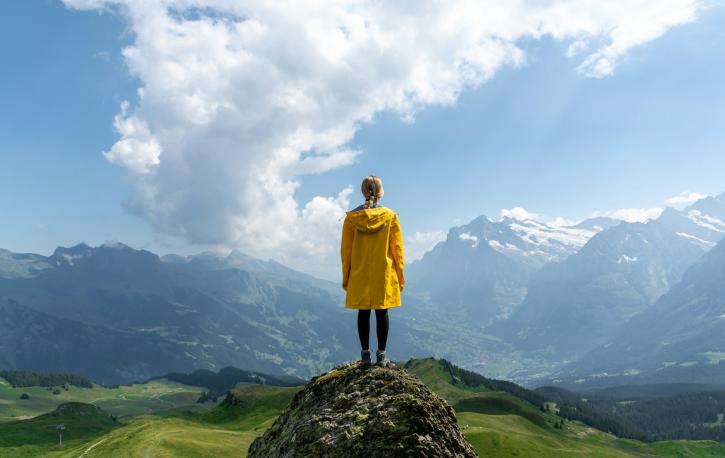 Rückansicht einer Frau in gelber Jacke, die von einer Anhöhe in eine bergige Landschaft schaut