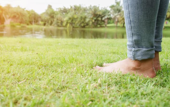 Man sieht zwei nackte Füße einer Person, die auf einer grünen Wiese steht, im Hintergrund ein See und Bäume.