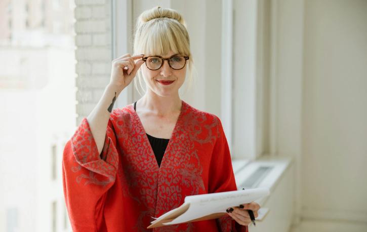 Eine blonde Frau mit Brille fasst sich an die Brille, sie hält einen Papierblock in der Hand.