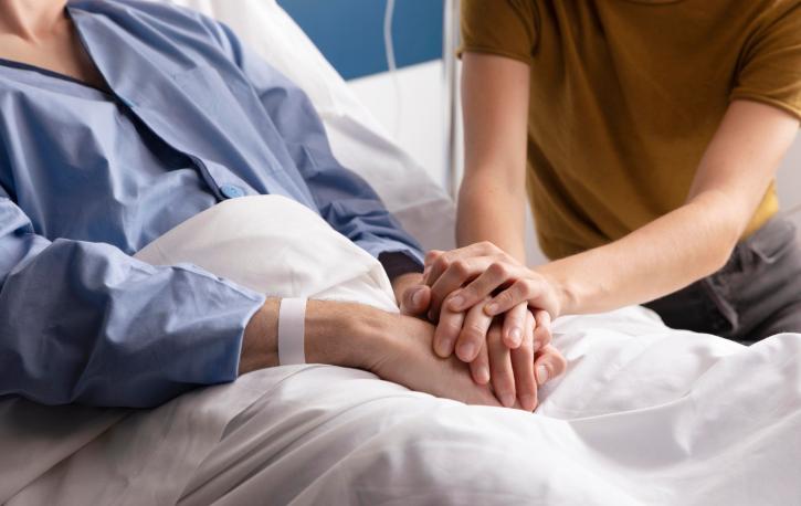 Eine junge Person hält ihre Hände auf die verschränkten Hände einer alten Person, die in einem Bett liegt.