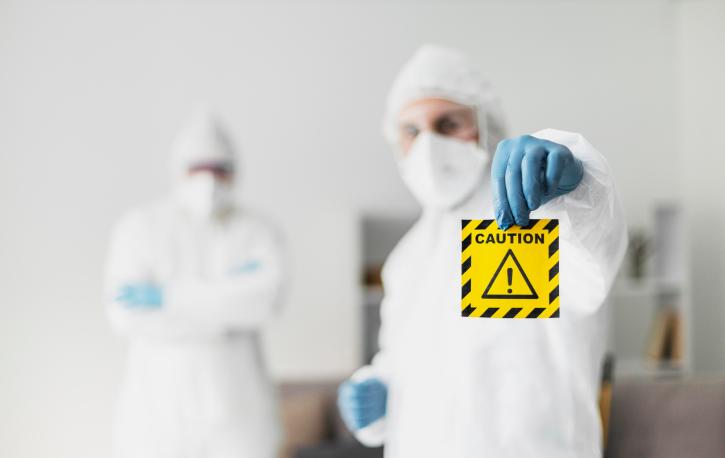 Zwei Personen in Hygieneschutzkleidung. Eine Person hält ein Achtung-Symbol hoch.