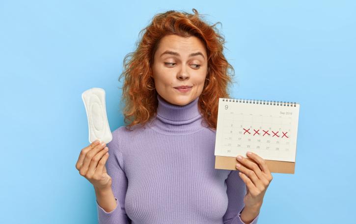 Eine Frau hält eine Binde und einen Zykluskalender in der Hand und schaut skeptisch.