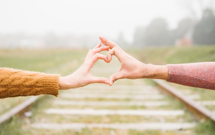 Die Hände von zwei Personen formen ein Herz, auf dem Boden sieht man Bahngleise.
